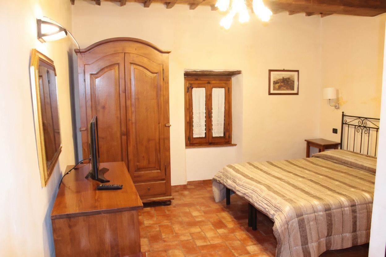 Cozy rustic bedroom at La Buca di Michelangelo hotel in Caprese Michelangelo, featuring a wooden wardrobe, wall mirror, flat-screen TV on a wooden dresser with remote, small window with lace curtains, framed artwork, bedside lamp, nightstand, double bed with striped bedspread and black metal headboard, terracotta tiled floor, and wooden beamed ceiling