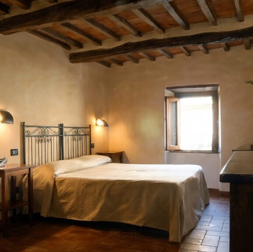 "Rustic double bedroom at La Buca di Michelangelo hotel in Caprese Michelangelo, featuring exposed wooden beam ceiling, wrought-iron headboard bed with beige striped bedspread, wooden nightstands with wall lamps, telephone, small window letting in natural light, framed artwork, terracotta tiled floor, and wooden desk."