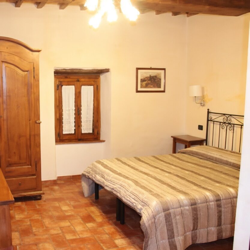 Cozy rustic bedroom at La Buca di Michelangelo hotel in Caprese Michelangelo, featuring a wooden wardrobe, wall mirror, flat-screen TV on a wooden dresser with remote, small window with lace curtains, framed artwork, bedside lamp, nightstand, double bed with striped bedspread and black metal headboard, terracotta tiled floor, and wooden beamed ceiling